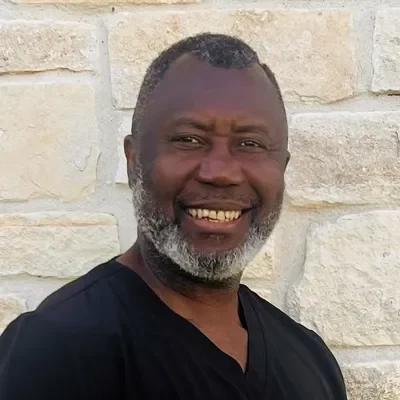 Dr. Roland Njumentoko smiling in front of a stone wall.